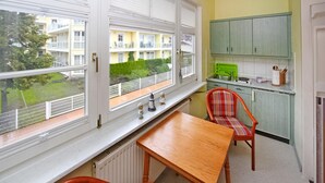 Et lille køkken med et bord og stole, et vindue der viser udsigt til en bygning og grøntområder, samt et grønt skab.