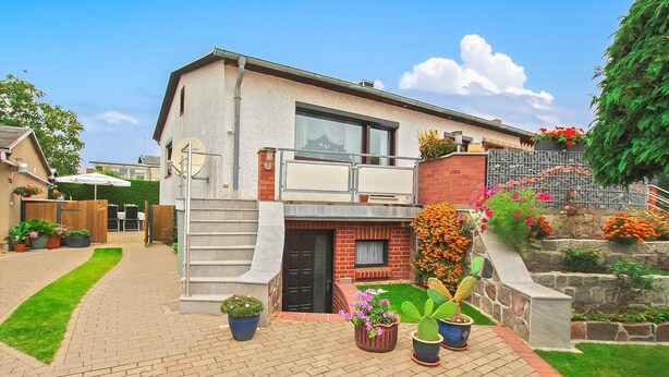 Une maison à deux étages avec un balcon, une entrée en briques et un jardin agrémenté de plantes en pot.