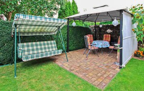 Ein überdachter Patio-Bereich mit Schaukelset, Tisch und Stühlen sowie ein kleiner Garten mit Blumen.