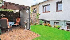 Un espace patio doté d’un coin salon couvert, d’une petite table et d’une chaise. On y trouve également un jardin avec des fleurs et une pelouse.