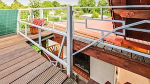 Um deck de madeira com guarda-corpo de metal, um vaso de planta e vista para um prédio com telhado vermelho.