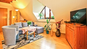Uma sala de estar aconchegante no estilo loft, com um sofá, poltrona e uma mesa pequena com um letreiro que diz 'Bem-vindo'. Há uma TV em um armário de madeira, uma janela grande com plantas e teto de madeira.