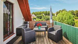 Uma varanda com móveis de vime, uma mesa com um vaso de flores e uma vista de vegetação e casas ao fundo.