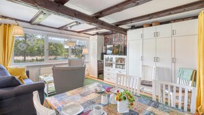 Ein gemütliches Zimmer mit einem großen Fenster, einem Esstisch gedeckt mit Tellern, Tassen und einer Blumenvase. Ein Sofa mit Kissen und ein Stuhl mit Karomuster sind ebenfalls zu sehen. Im Hintergrund sind weiße Schränke mit Glastüren erkennbar.