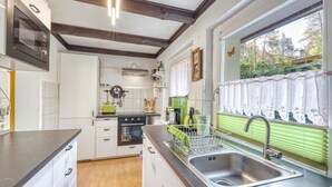 Eine Küche mit weißen Schränken, einer schwarzen Arbeitsplatte, einer Edelstahl-Spüle, einem grünen Wasserkocher und einem Fenster mit Spitzenvorhängen.
