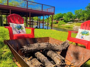 Een rustieke buitenzitplaats met een metalen vuurkorf en rode stoelen, met uitzicht op een rivier en een houten terras.