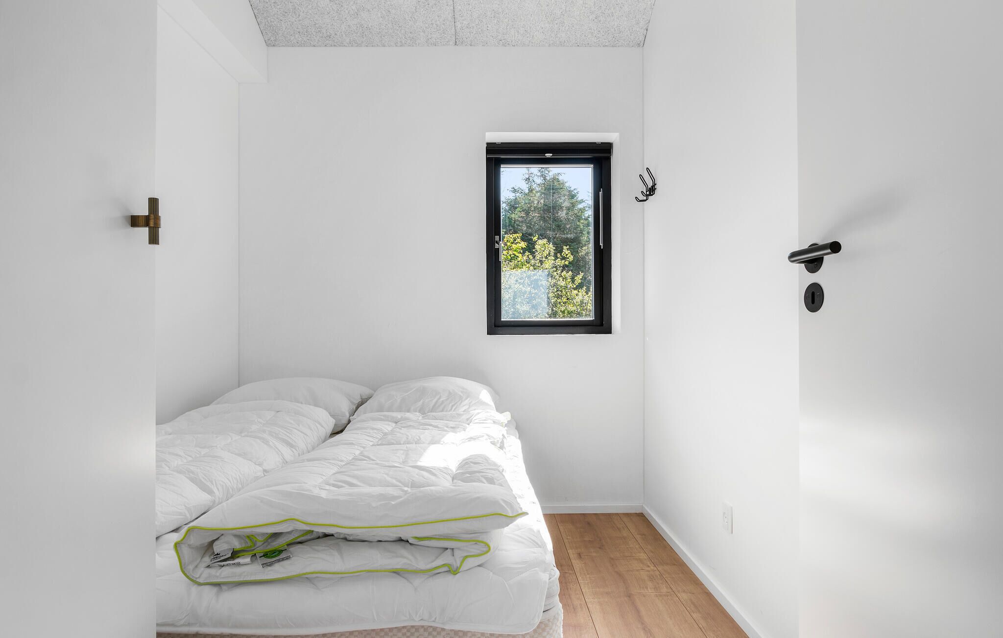 Une petite pièce lumineuse, avec un lit simple, une fenêtre donnant sur des arbres et une porte dotée d’une serrure.