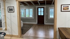 Ein geräumiger Raum mit Holzboden, Holzbalkendecke und einer Tür zu einem anderen Raum. An den Wänden befinden sich gerahmte Bilder und ein Fenster mit Blick ins Freie.