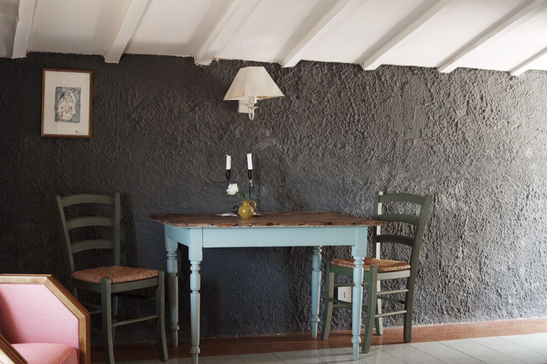 A room with a rustic stone wall, a wooden table with two chairs, a framed picture, and a wall-mounted lamp.