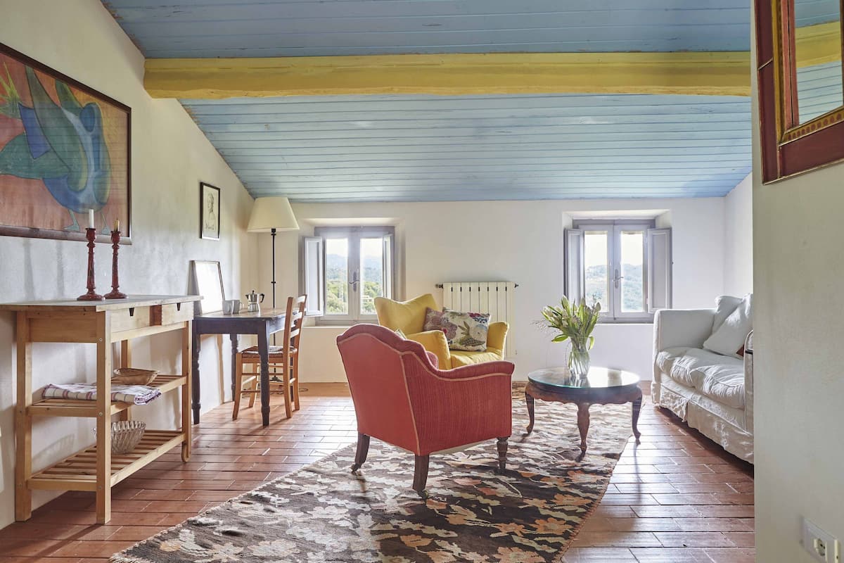 A cozy living room with a red armchair, a yellow armchair, and a white sofa. There is a wooden table with a lamp, a bookshelf, and a patterned rug.