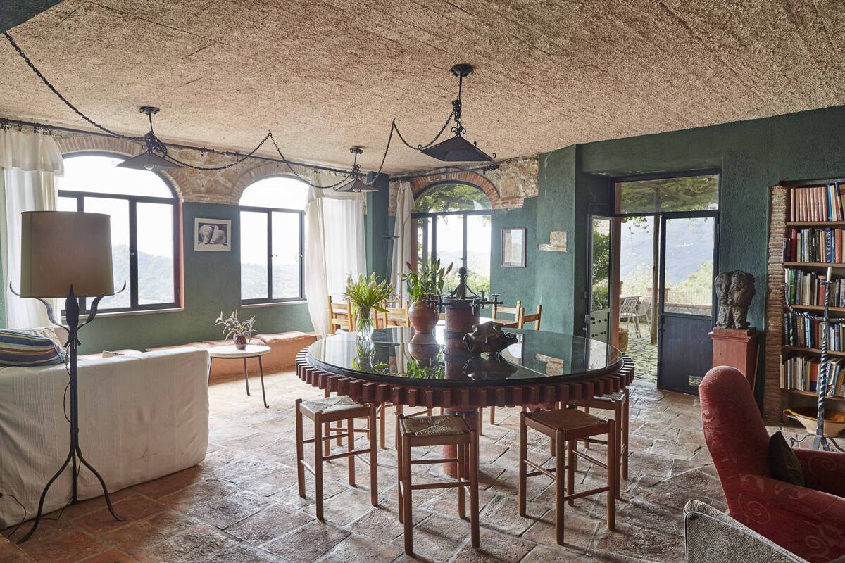 A dining area with a round table, chairs, and a chandelier. There are windows with views of greenery and a bookshelf with books.