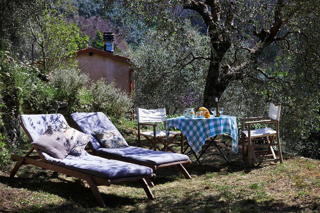 Un jardin avec deux chaises longues, une table recouverte d’une nappe à carreaux et, au fond, un bâtiment au style rustique.