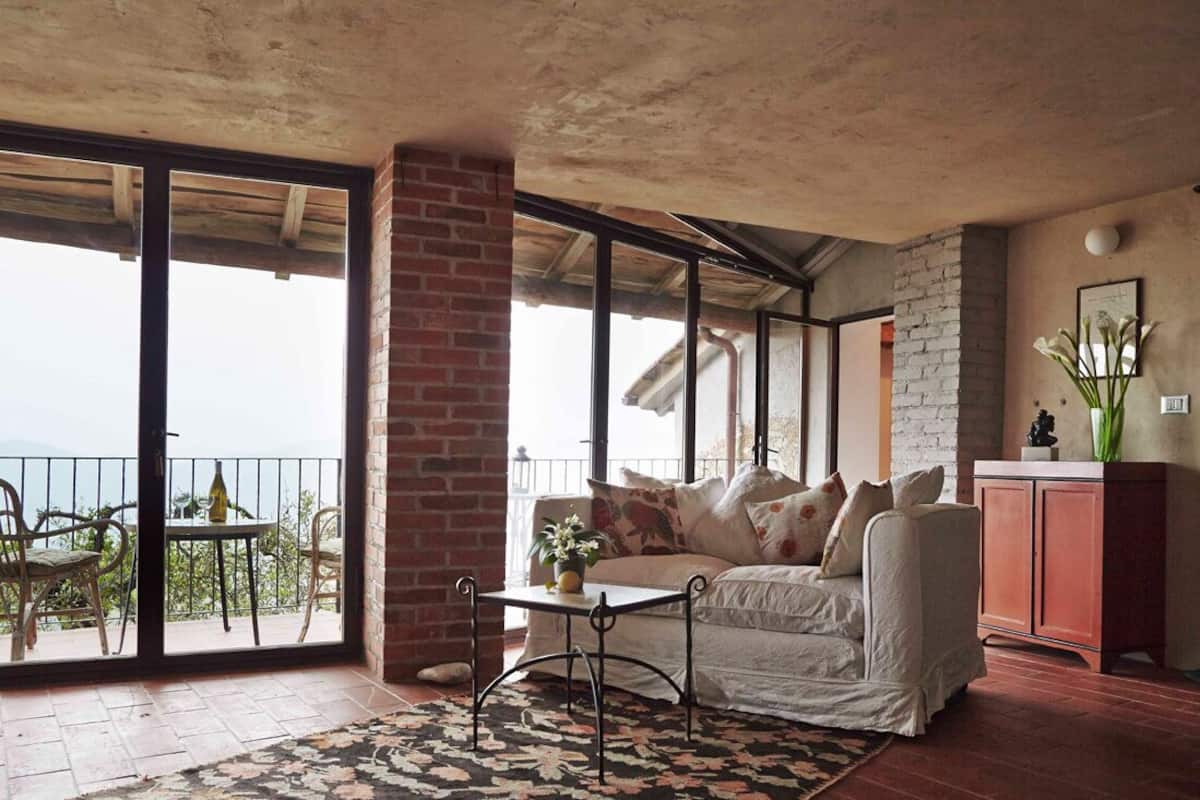 A cozy living room with a sofa, a small table, and a cabinet. There is a balcony with a view of the sky and a railing.