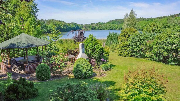 A gazebo with a brick base and a green roof, surrounded by lush greenery and a view of a lake.