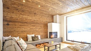 Een knusse woonkamer met een houten plafond, een open haard, een groot raam met uitzicht en een comfortabele bank.