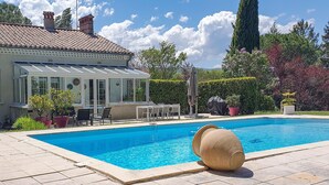 A swimming pool with a tiled deck, a covered patio with outdoor furniture, and a house with a tiled roof and large windows. - Stunning home in Tabre (Tabre)