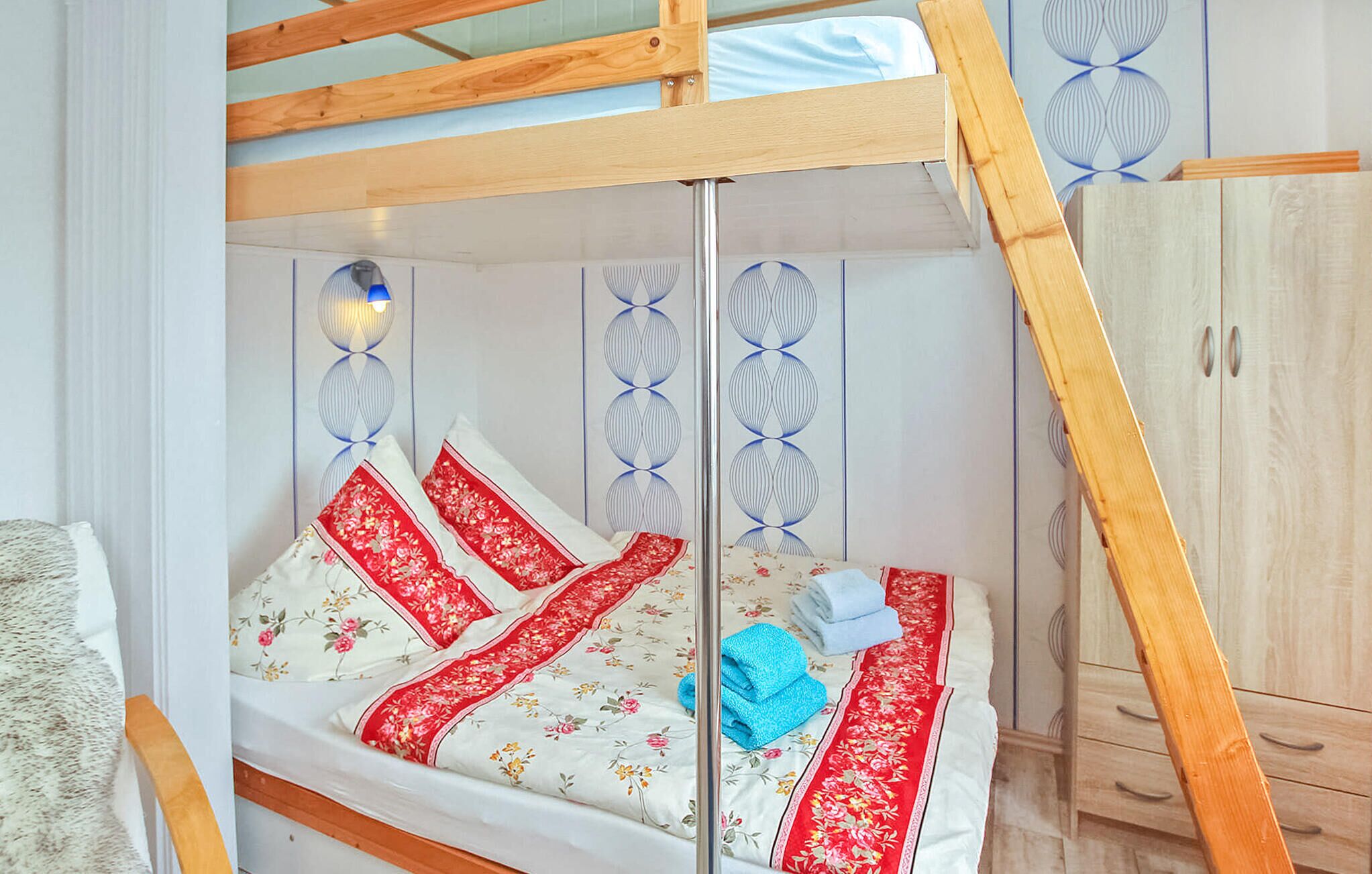 A bunk bed with a wooden frame, a white mattress with floral patterns, and red accents. There is a wooden wardrobe to the right and a wall with decorative panels.