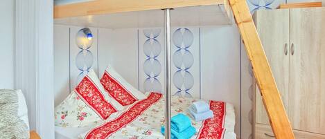 A bunk bed with a wooden frame, a white mattress with floral patterns, and red accents. There is a wooden wardrobe to the right and a wall with decorative panels.