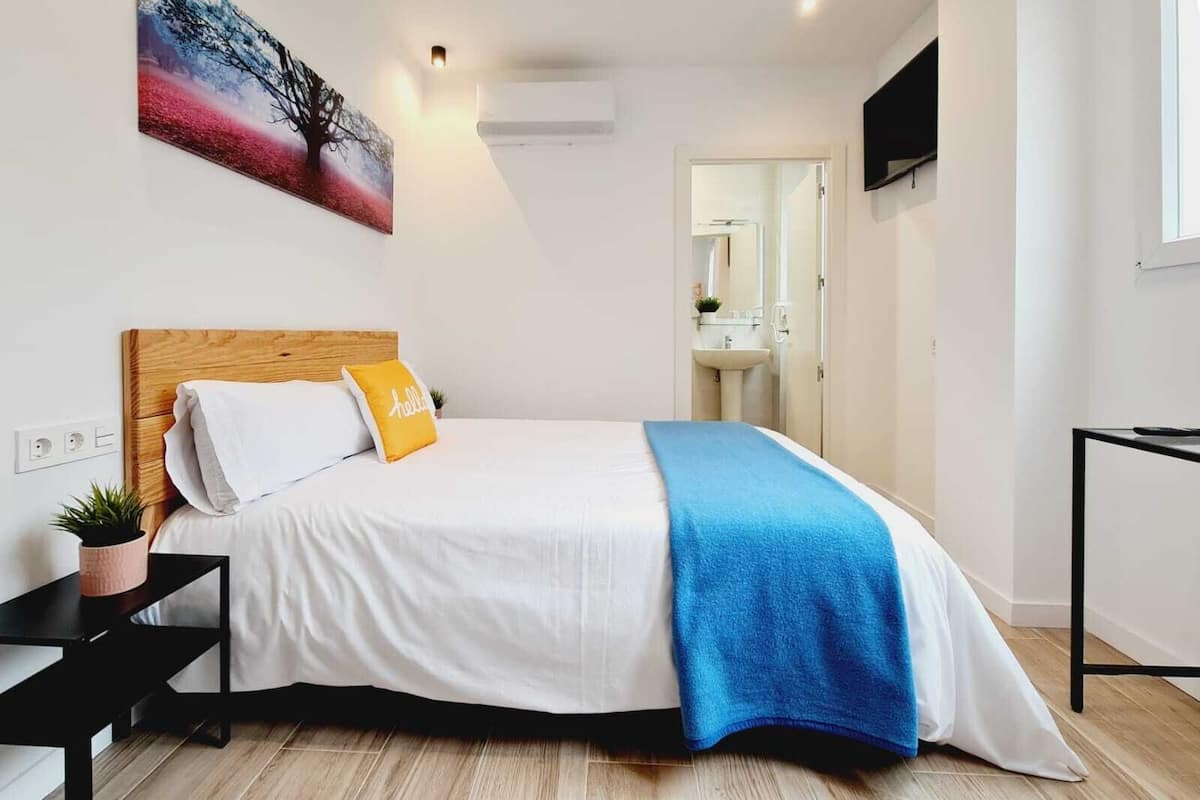 A modern bedroom with a wooden headboard, white bedding, a blue throw blanket, and a small plant on a bedside table.