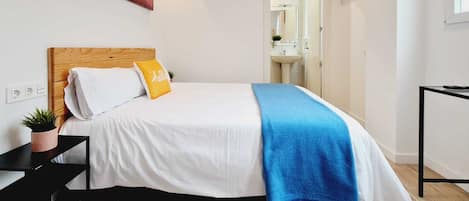 A modern bedroom with a wooden headboard, white bedding, a blue throw blanket, and a small plant on a bedside table.