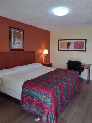 Una habitación de hotel con una cama grande, un escritorio, una silla, una mesita de noche con una lámpara y dos cuadros en las paredes.