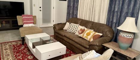 A living room with a brown sofa, beige armchair, and a white coffee table. There is a patterned rug on the floor, a door, and a painting on the wall.