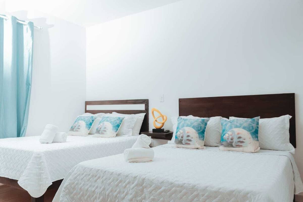 Dos camas individuales con ropa blanca y cabeceros a juego, una cortina azul y una mesita con un objeto decorativo.