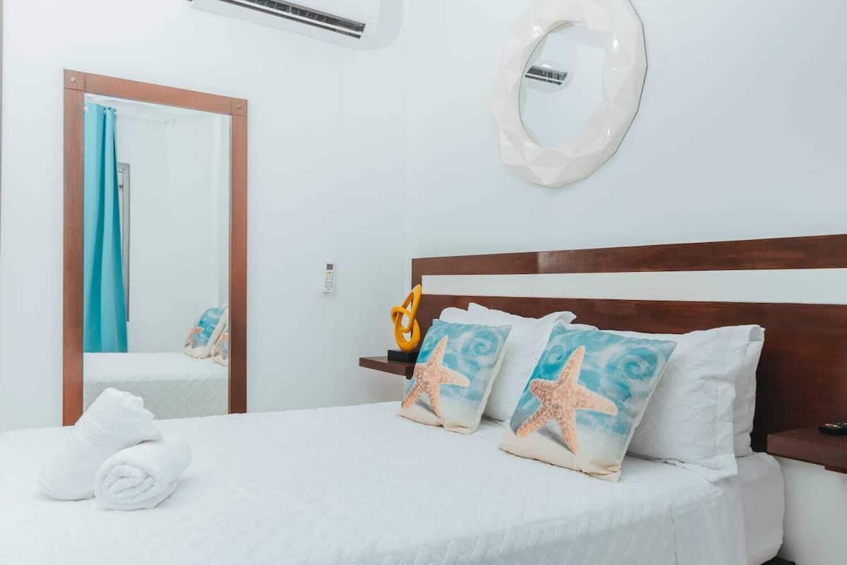 Una habitación de hotel con una cama, cabecero de madera, aire acondicionado montado en la pared, un espejo y una toalla sobre la cama.