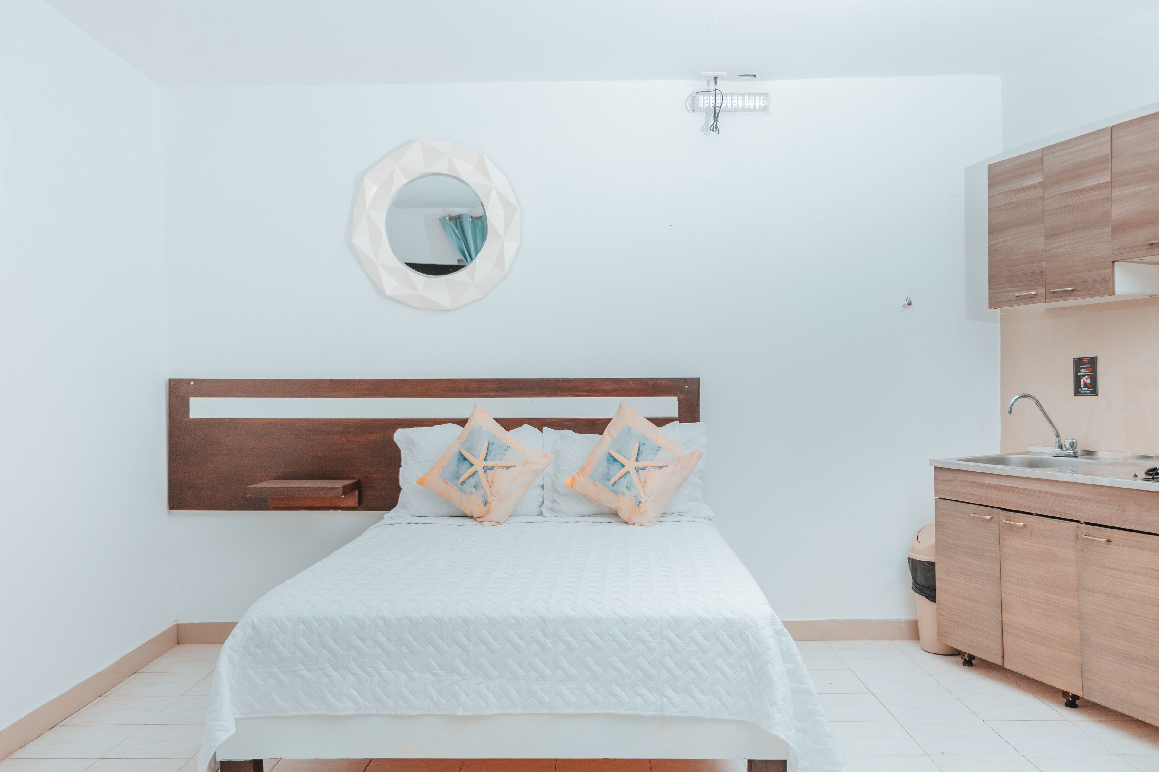 A bedroom with a bed, a bedside table, a mirror, and a kitchenette with cabinets and a sink.