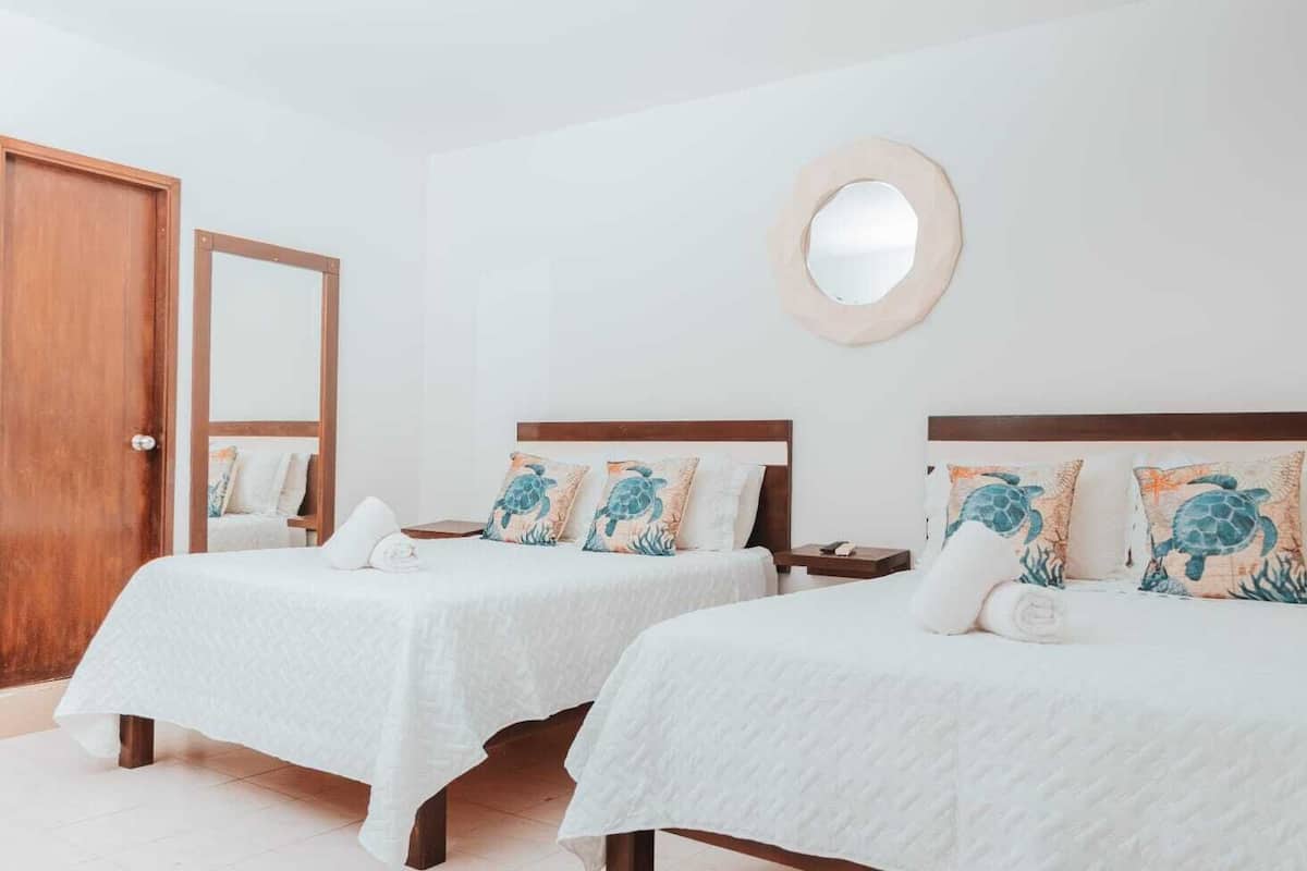 Una habitación de hotel con dos camas, una puerta de madera, un espejo redondo y un suelo de baldosas blancas.