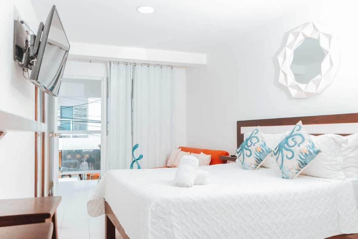 Habitación de hotel con cama, televisor montado en la pared, balcón con barandilla y vistas al exterior.