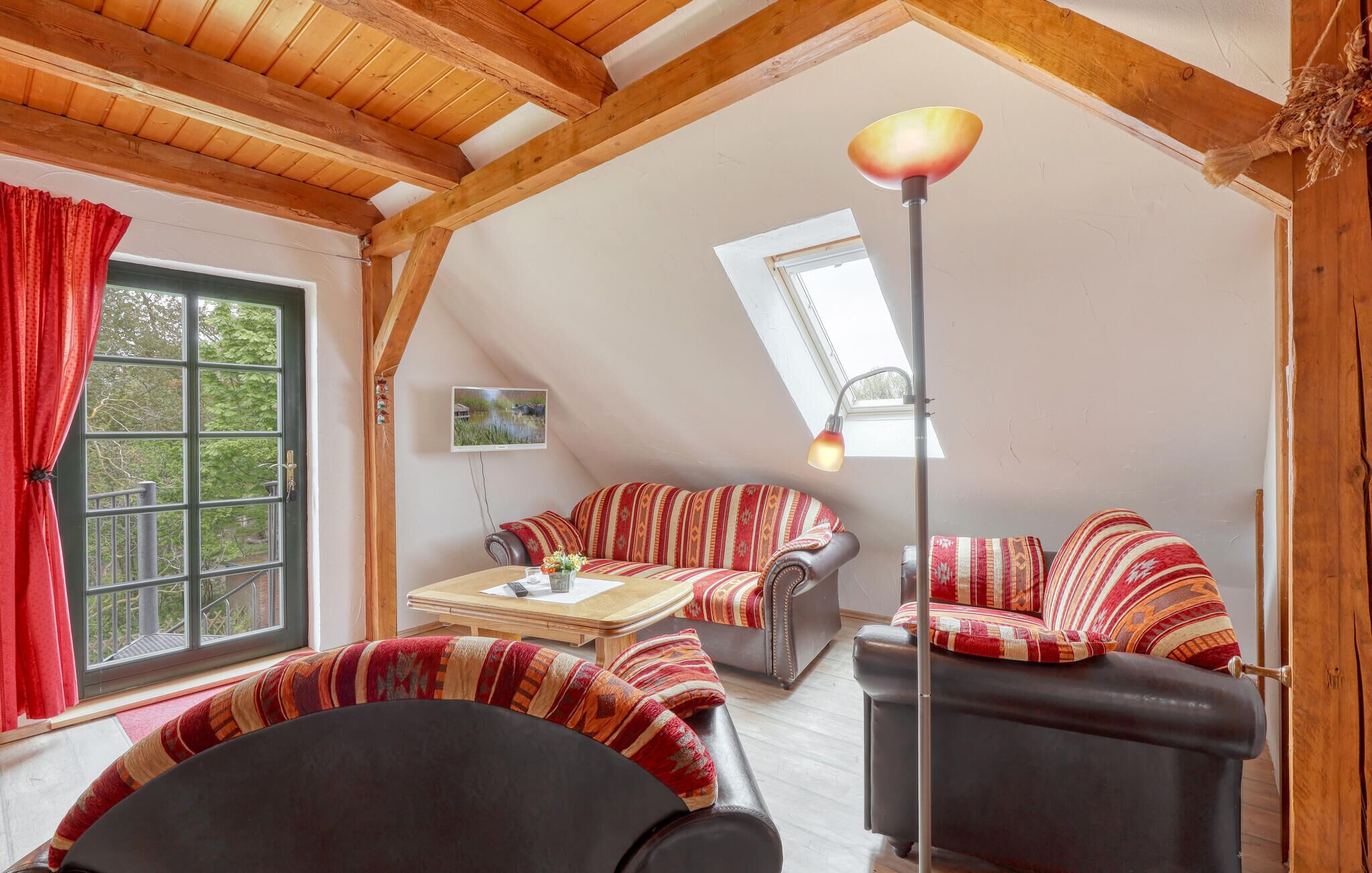 A cozy loft-style living room with a high ceiling, wooden beams, a large window with red curtains, a sofa set with patterned cushions, a coffee table, and a floor lamp.