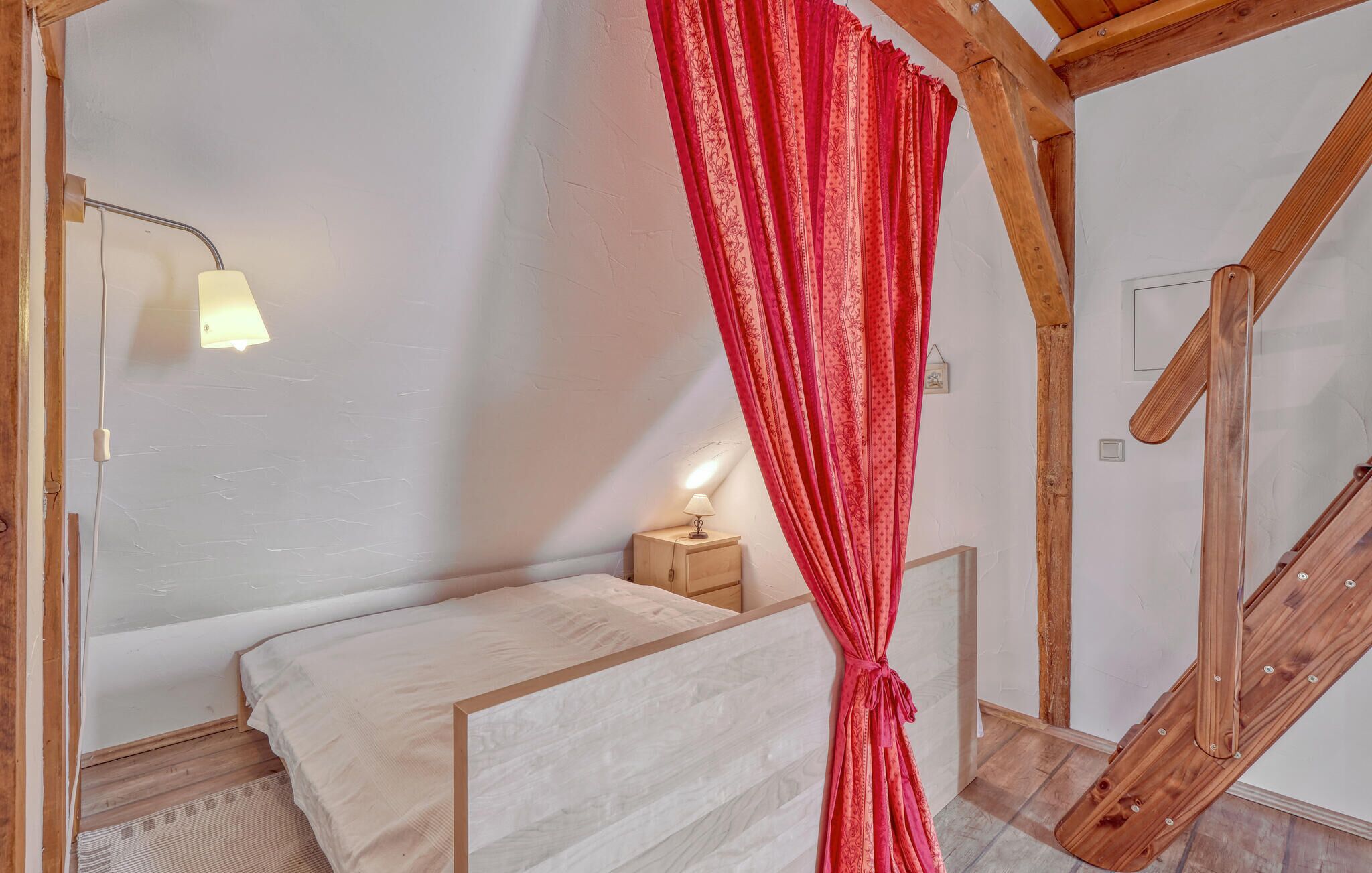 Ein Schlafzimmer mit einem Bett, einem Nachttisch, einer Lampe, einem roten Vorhang und einer Holztreppe.