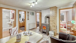 A cozy living room with a fireplace, a dining area with a table set for four, and a kitchen visible through an open door. - Ferienhaus Rheinsberg OT Kagar (Rheinsberg OT Kagar)