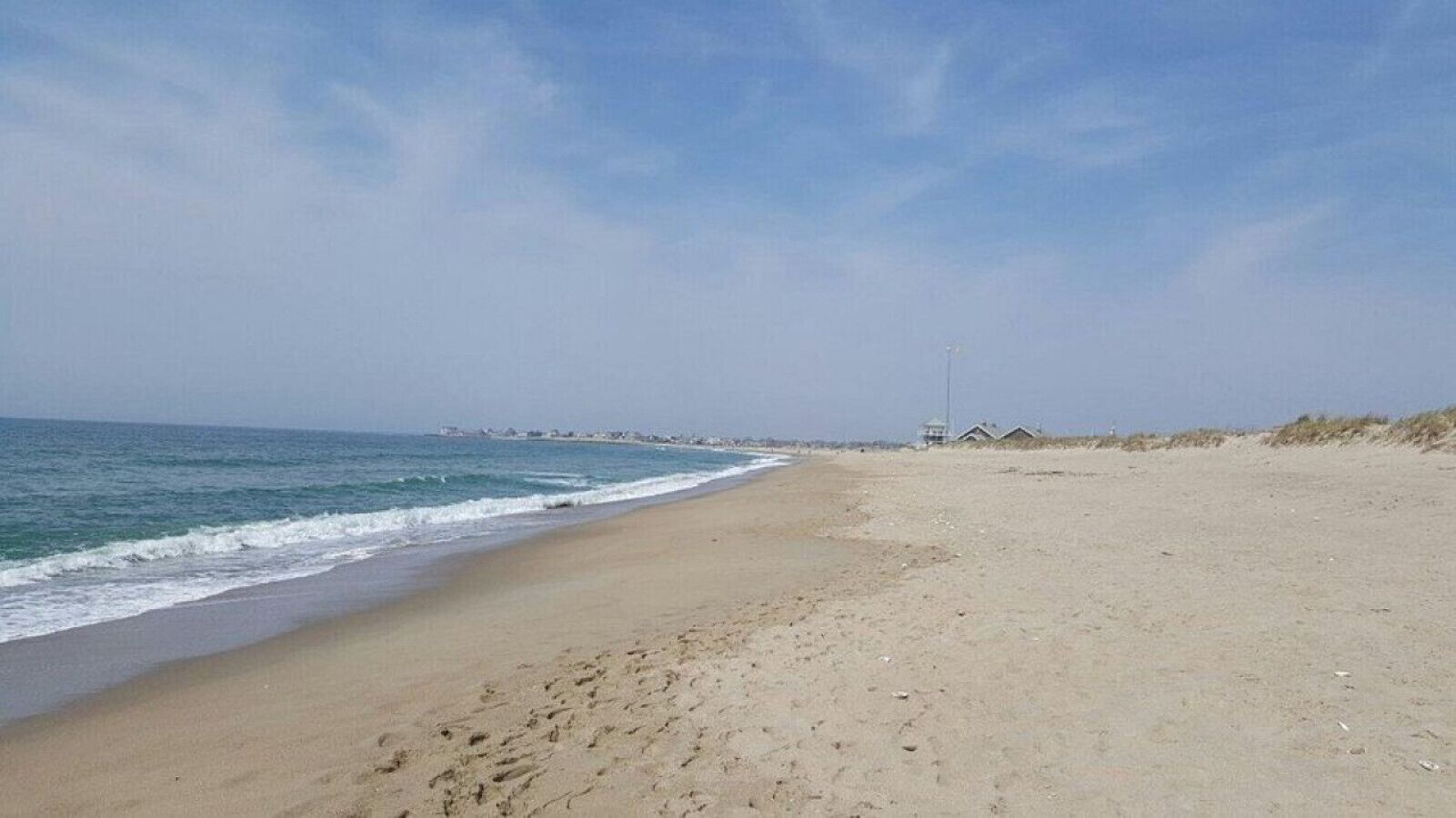 Uma praia extensa e arenosa, com ondas suaves, céu claro e alguns edifícios ao longe.