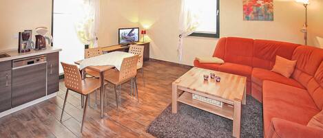 A modern living room with a wooden floor, a red sofa, a coffee table, a dining table with chairs, a TV, and a kitchen area with a coffee maker and cabinets.