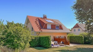 Une maison à deux étages avec un toit recouvert de tuiles rouges, un petit balcon et une cour arrière clôturée, aménagée avec du mobilier d’extérieur.