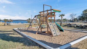 A wooden playground with a slide and climbing wall, surrounded by a sandy area and a body of water in the background.