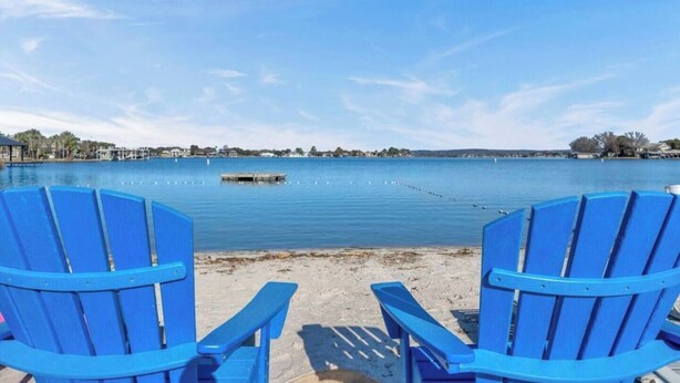 Two blue Adirondack chairs facing a calm lake with a dock and buildings in the background.