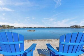 Zwei blaue Adirondack-Stühle, die auf einen ruhigen See mit einem Steg und Gebäuden im Hintergrund blicken.