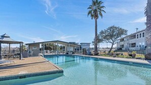 A clear swimming pool with a covered area, palm trees, and a building with a flag.