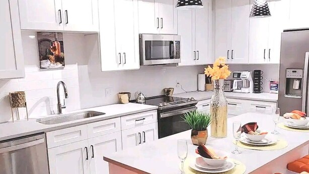 A modern kitchen with white cabinets, a stainless steel sink, a microwave, and a stove. There are orange chairs around the island, and a vase with flowers on the counter. - Lux and Cozy 2beds 2 Baths up to 8 guest (West Palm Beach)