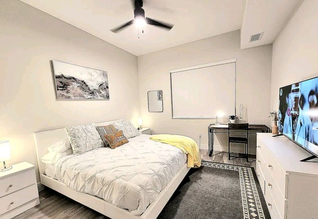 A bedroom with a bed, a desk, a television, and a ceiling fan.