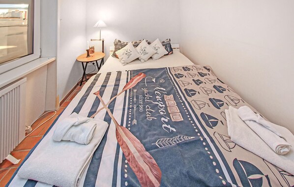A neatly made bed with a striped bedspread and decorative pillows, a small bedside table with a lamp, and a window with a view. - Ferienwohnung Prerow Ostseebad (Prerow (Ostseebad))