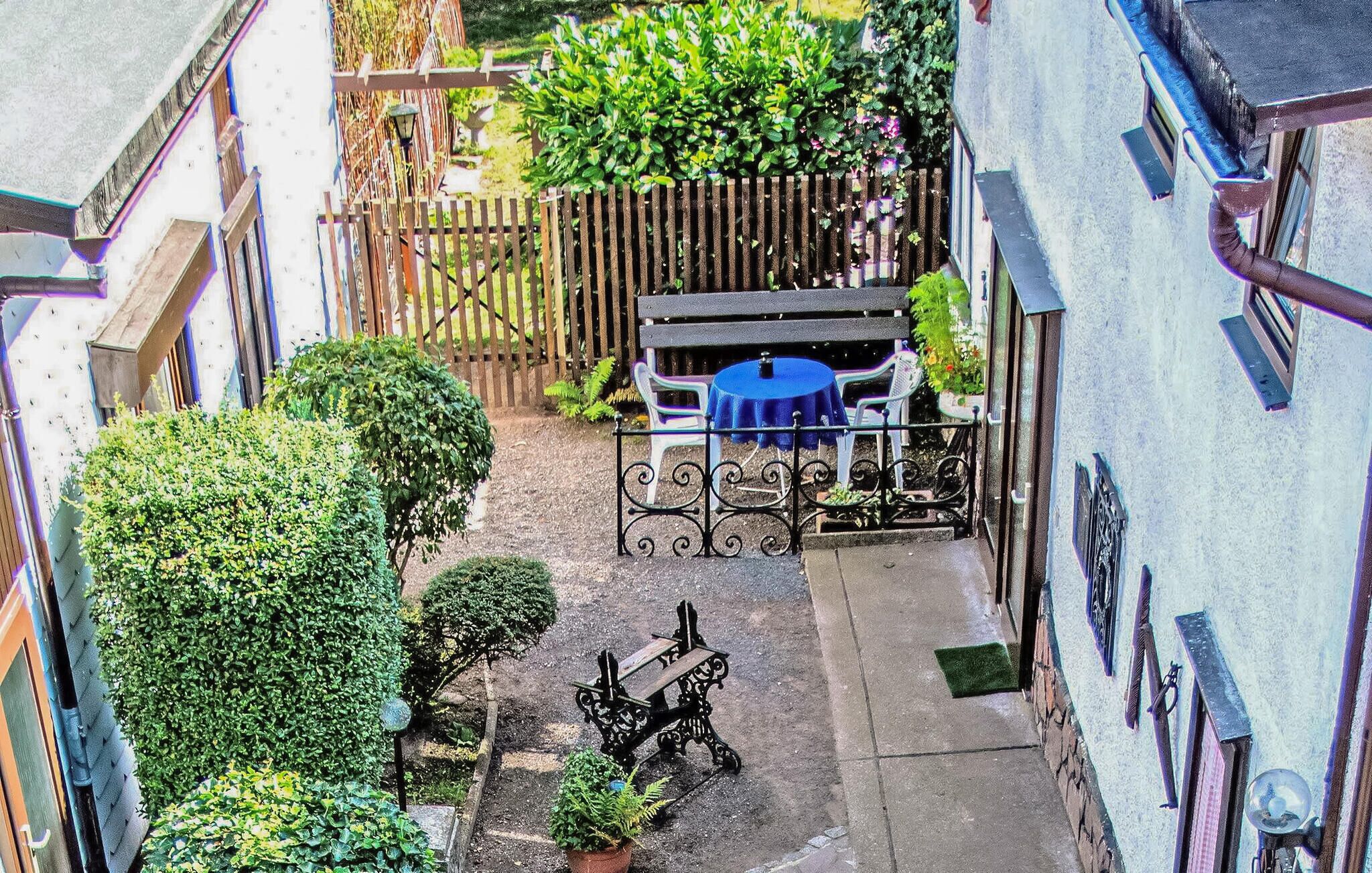 A narrow alleyway with a small courtyard featuring a table and chairs, surrounded by greenery and a wooden fence.