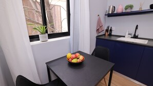 Et lille køkken med spisebord, skålsæt frugt og en plante ved vinduet.