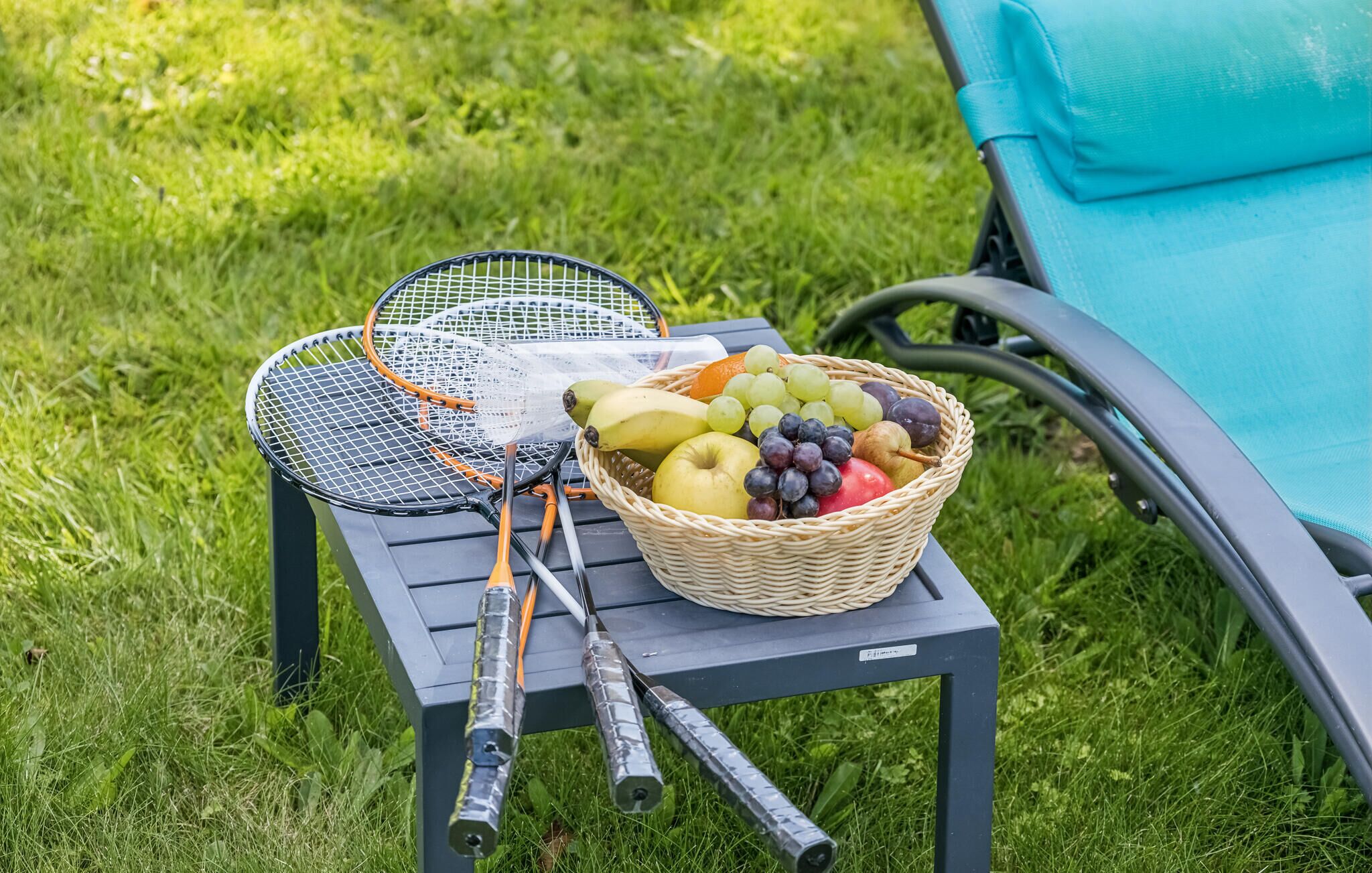 Ein Tisch mit Badmintonschlägern, einer Fruchtkorb und ein Stuhl in einer grünen Außenanlage.