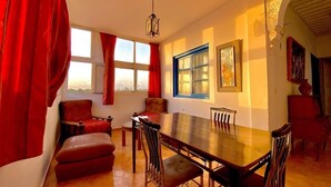 A dining area with a long wooden table, red curtains, and a view of the cityscape through the windows. - Riad Dar Baddi Avec 9 Suite : Marrakech (Marrakech)