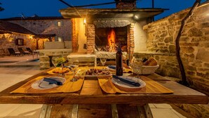 Uma área de refeições ao ar livre com parede de pedra, forno de tijolos e uma mesa posta com pratos, copos e garrafa.