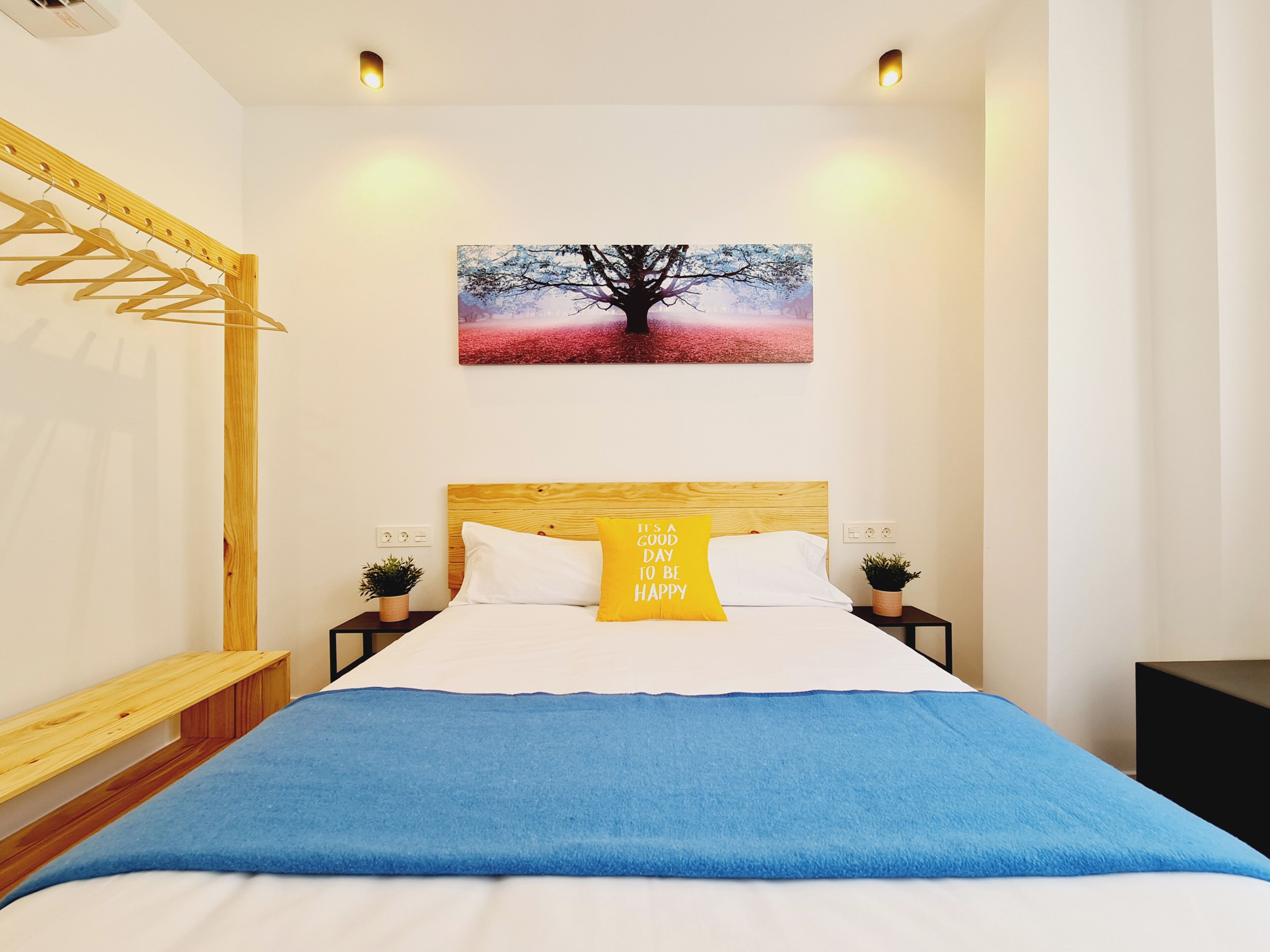 Ein Schlafzimmer mit Holz-Kopfteil, einem Bett mit weißer und blauer Bettwäsche, einem gelben Kopfkissen mit Motivationsspruch, einem an der Wand hängenden Baumgemälde und einem Holz-Kleiderständer.
